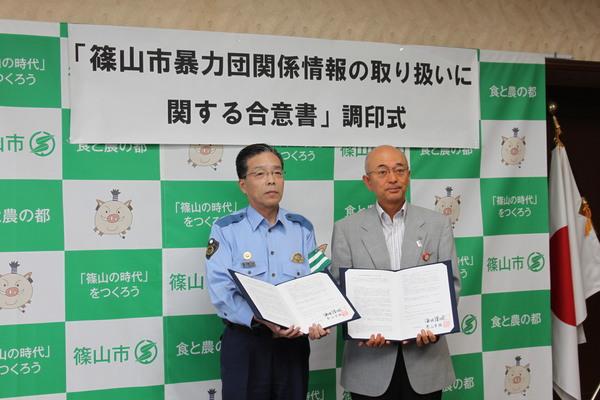 左側に警察署長、右側に市長が並んで立ち調印した合意書を見開き両手に持って立っている写真